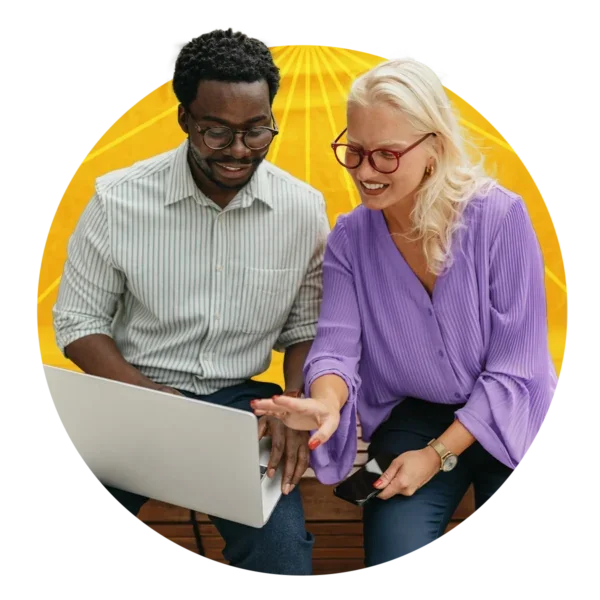 Two people wearing button downs and glasses sit beside each other and study the screen of an open laptop.