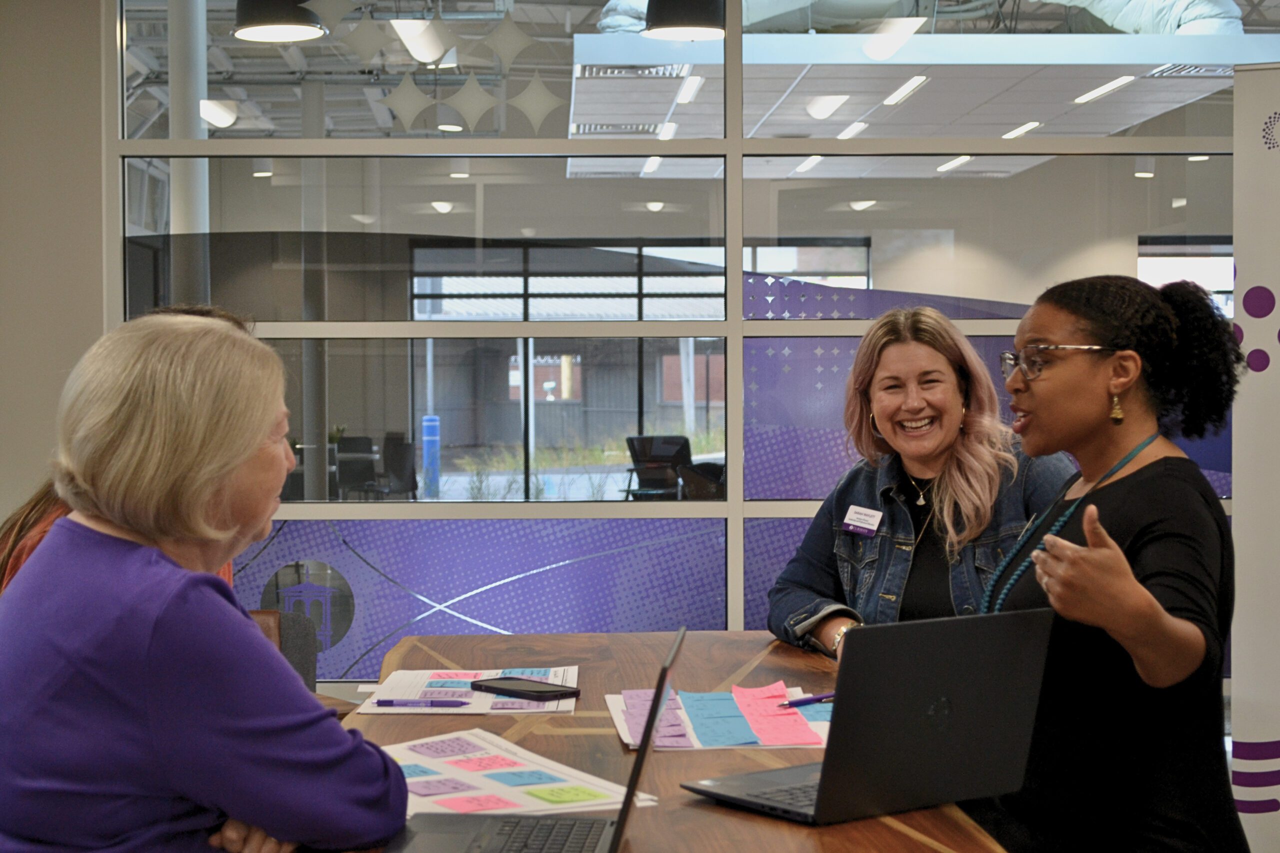 Three professionals speak to each other at a table that contains multiple documents with sticky notes and an open laptop.
