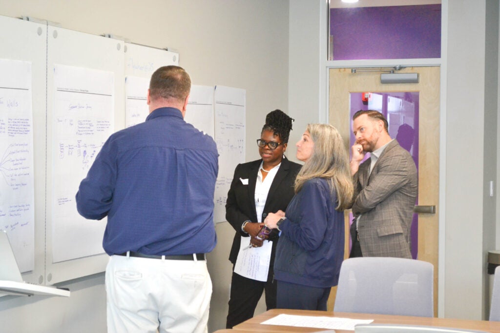 A group of diverse leaders are gathered around discussing hand-drawing planning posters on the wall.